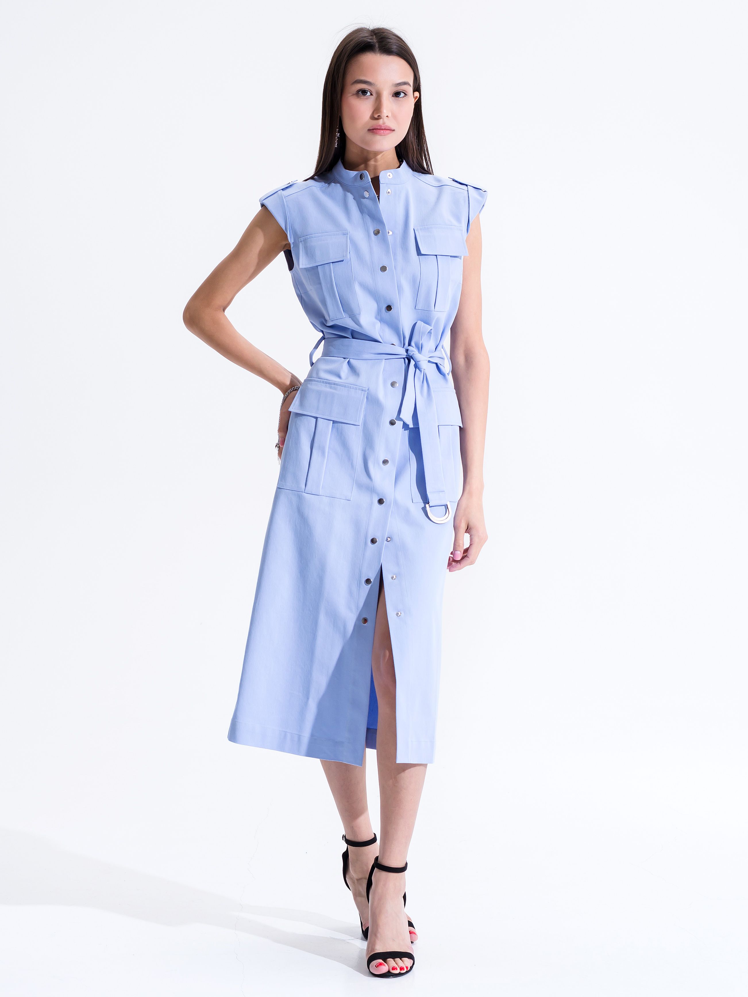 Woman wearing a light blue utility midi shirt dress with a band collar, cap sleeves, chest flap pockets, self-tie belt, bellows hip pockets, and a buttoned front slit on a clean white background